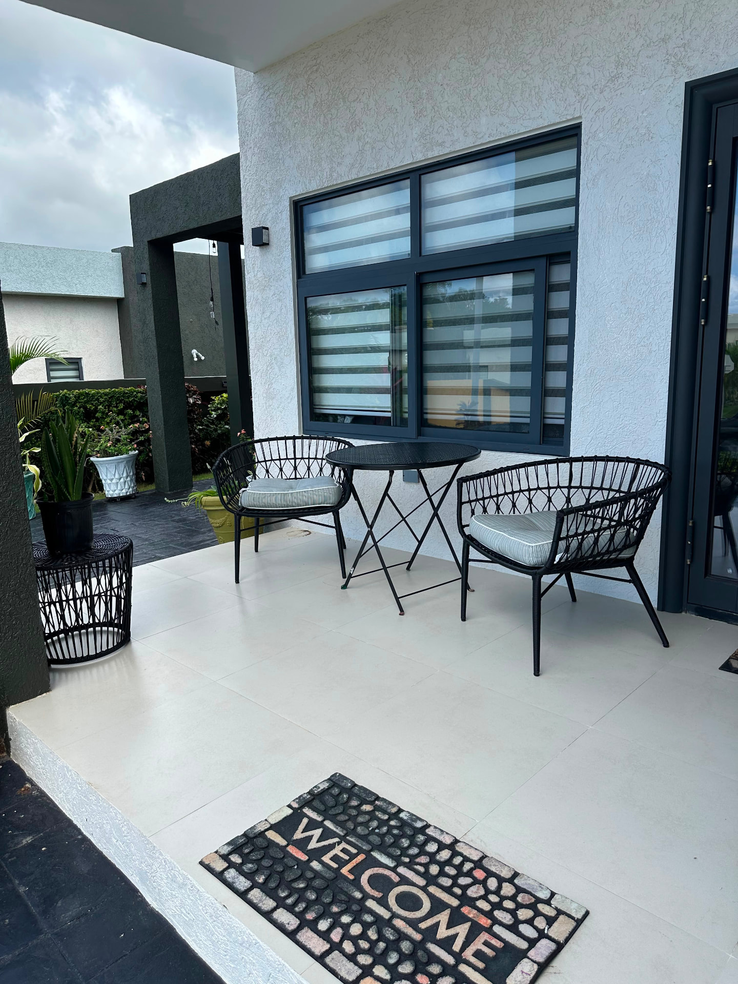 A covered patio with rattan chairs and a table, a hand-crafted welcome mat at the door